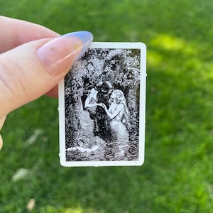 May include: A small, rectangular card featuring a black and white illustration of two figures in a forest setting. One figure appears to be a mermaid. The card is held against a backdrop of green grass.