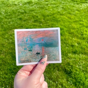 Op de afbeelding: Een sticker met een schilderij van een zonsopgang boven water met een klein bootje op de voorgrond. Het schilderij is in een gedempte kleurenpalet van roze, oranje, blauw en grijs.