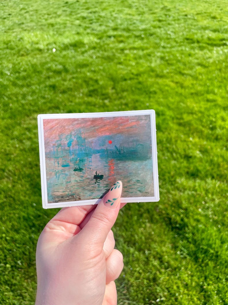 Op de afbeelding: Een sticker met een schilderij van een zonsopgang boven water met een klein bootje op de voorgrond. Het schilderij is in een gedempte kleurenpalet van roze, oranje, blauw en grijs.