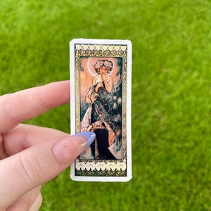 May include: A sticker featuring a vintage illustration of a woman standing in a forest with a crescent moon behind her. The woman is wearing a long, flowing dress with a floral pattern. The sticker has a white background with a gold border.