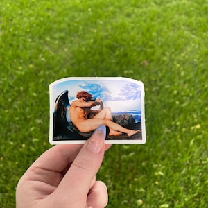 Puede incluir: Una pegatina con una pintura de una figura alada con alas azules y blancas, sentada en una roca con un cielo azul y nubes en el fondo.