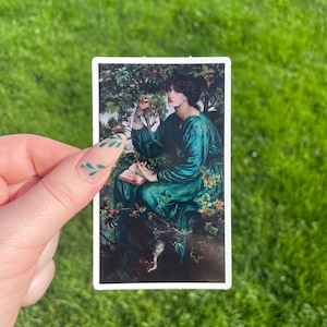 May include: A sticker featuring a painting of a woman in a green dress sitting in a forest setting. She is holding a book and looking at a small bird.