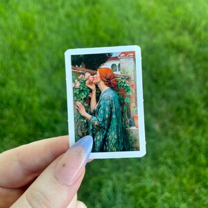 May include: A sticker featuring a painting of a woman in a green dress smelling a pink rose. The woman has red hair and is standing in a garden setting.