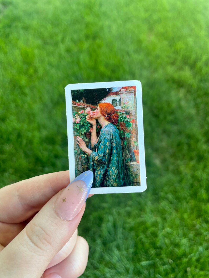 May include: A sticker featuring a painting of a woman in a green dress smelling a pink rose. The woman has red hair and is standing in a garden setting.