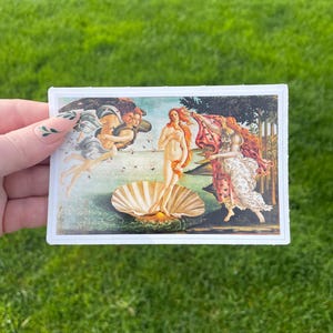 May include: A vintage-style print of the famous Renaissance painting "The Birth of Venus" by Sandro Botticelli. The painting depicts a nude woman standing on a giant scallop shell, with the wind blowing her hair and a flowing drapery.