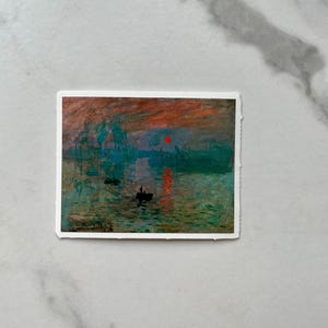 Op de afbeelding: Een schilderij van een zeegezicht met een rode zon die ondergaat boven het water. Het water is een mix van blauw, groen en grijs, en er zijn verschillende boten in de verte. Het schilderij is ingelijst in een witte lijst.