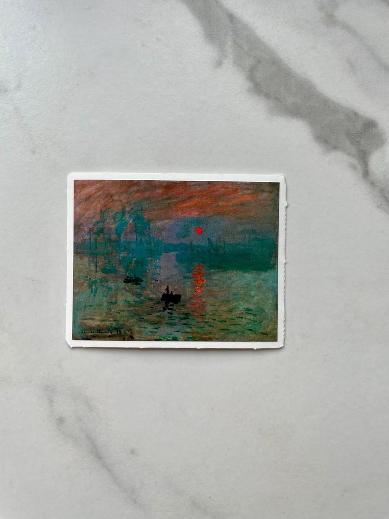 Op de afbeelding: Een schilderij van een zeegezicht met een rode zon die ondergaat boven het water. Het water is een mix van blauw, groen en grijs, en er zijn verschillende boten in de verte. Het schilderij is ingelijst in een witte lijst.