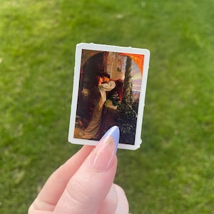 May include: A vintage sticker featuring a painting of a couple embracing in a romantic setting. The painting is set against a backdrop of a stone building and a lush garden. The couple is dressed in period clothing and the colors are warm and inviting.