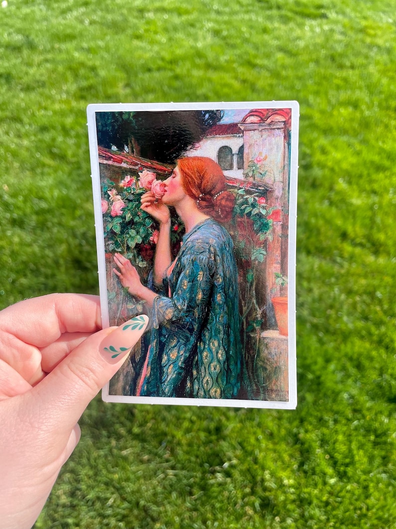 May include: A painting of a woman with red hair smelling a pink rose. She is wearing a blue and gold patterned dress and is standing in a garden setting.