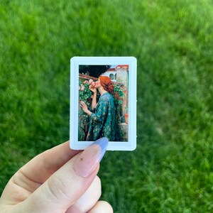 May include: A sticker featuring a painting of a woman with red hair smelling a pink rose. The woman is wearing a green and gold patterned dress. The background is a garden with a building in the distance.