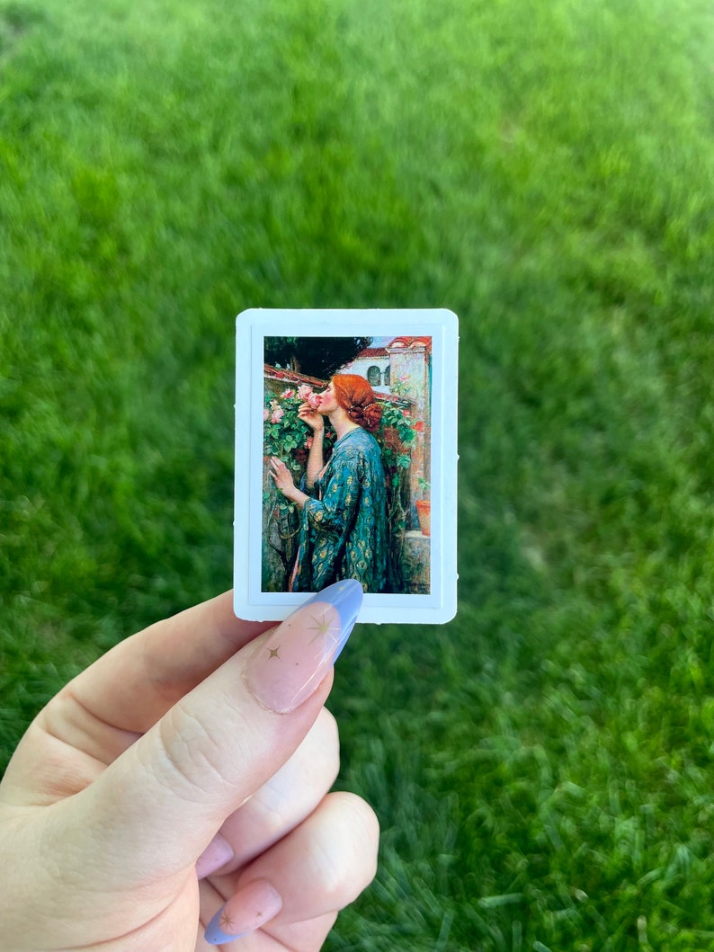 May include: A sticker featuring a painting of a woman with red hair smelling a pink rose. The woman is wearing a green and gold patterned dress. The background is a garden with a building in the distance.