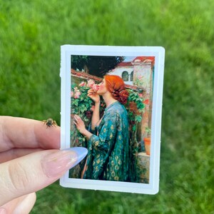 May include: A sticker featuring a painting of a woman with red hair smelling a pink rose. The woman is wearing a blue and gold patterned robe. The background is a garden with a building in the distance.