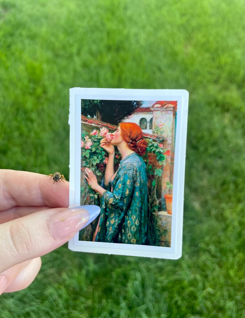May include: A sticker featuring a painting of a woman with red hair smelling a pink rose. The woman is wearing a blue and gold patterned robe. The background is a garden with a building in the distance.