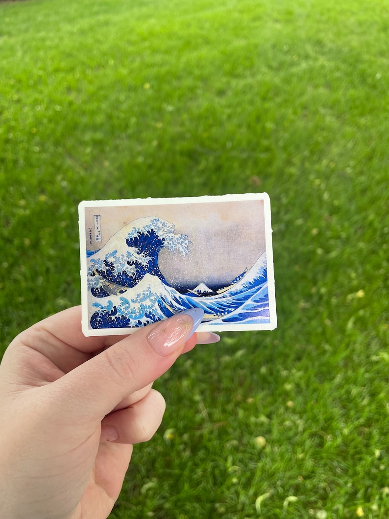 Op de afbeelding: Een sticker met een blauwe en witte illustratie van een grote golf die tegen een berg aan beukt. De golf is afgebeeld in een traditionele Japanse stijl, met een gevoel van beweging en energie.