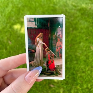 May include: A rectangular sticker depicting a painting of a medieval scene. A woman in a white gown is giving a sword to a kneeling knight in red armour. Other figures stand in the background. The sticker is held against a green grassy background.