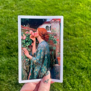 May include: A sticker featuring a painting of a woman in a blue and gold robe smelling a pink rose. The woman has red hair and is standing in a garden setting.