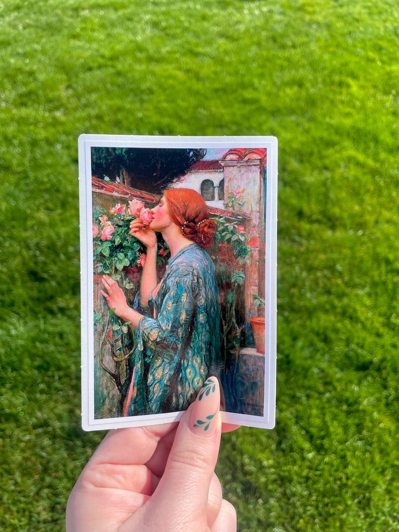 May include: A sticker featuring a painting of a woman in a blue and gold robe smelling a pink rose. The woman has red hair and is standing in a garden setting.