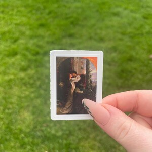 May include: A sticker featuring a painting of a woman in a white dress standing in a doorway with a man behind her. The painting is set against a backdrop of a castle and a sunset.