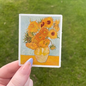 Puede incluir: Una pegatina con una impresión de la pintura "Girasoles" de Van Gogh. La obra representa un ramo de girasoles amarillos en un jarrón amarillo, sobre un fondo azul claro. El nombre del artista, "Vincent", es visible en el jarrón.