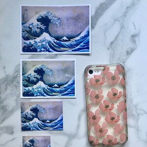 Op de afbeelding: Vier verschillende formaten van Great Wave off Kanagawa-prints, een beroemde Japanse houtsnede van Hokusai, worden getoond op een wit marmeren oppervlak. Een transparante telefoonhoes met een roze bloemenpatroon is ook te zien.