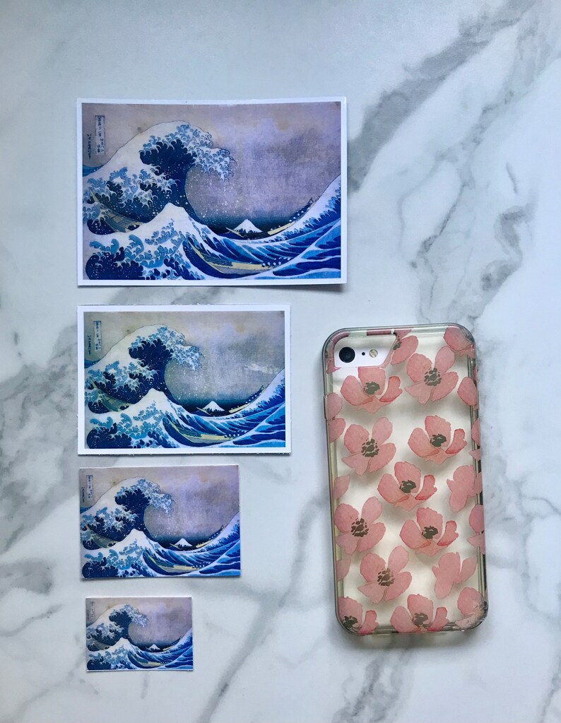 Op de afbeelding: Vier verschillende formaten van Great Wave off Kanagawa-prints, een beroemde Japanse houtsnede van Hokusai, worden getoond op een wit marmeren oppervlak. Een transparante telefoonhoes met een roze bloemenpatroon is ook te zien.