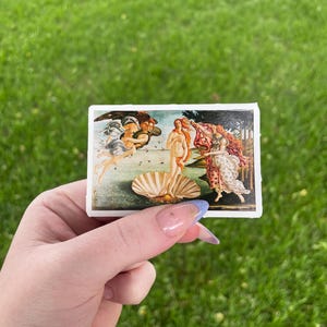 May include: A sticker featuring a reproduction of Sandro Botticelli's The Birth of Venus painting. The painting depicts a nude woman standing on a giant scallop shell, with the wind god Zephyrus and the nymph Chloris blowing her towards shore.