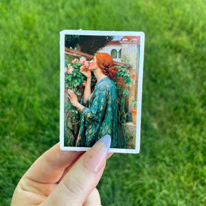 May include: A sticker featuring a painting of a woman with red hair smelling a pink rose. The woman is wearing a green and gold patterned robe. The background is a garden with a building in the distance.