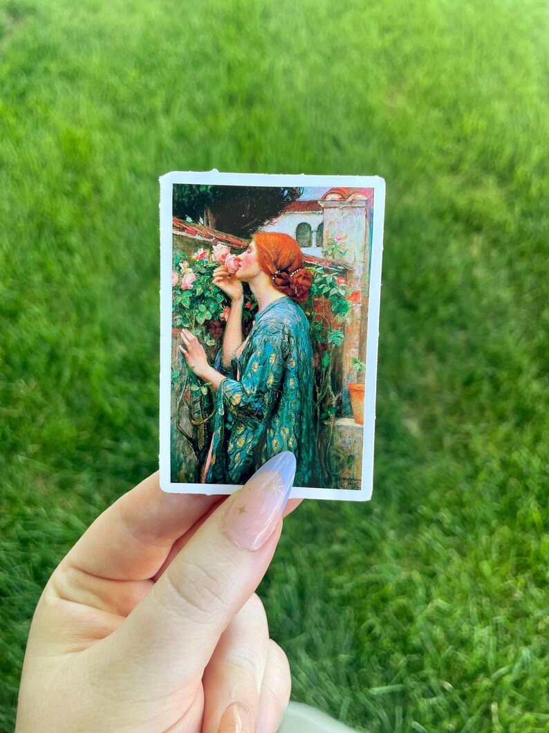 May include: A sticker featuring a painting of a woman with red hair smelling a pink rose. The woman is wearing a green and gold patterned robe. The background is a garden with a building in the distance.