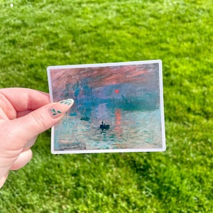 Op de afbeelding: Een sticker met een schilderij van een zonsopgang boven water met een klein bootje op de voorgrond. De lucht is een mix van roze, oranje en paars, en het water is diepblauw. Het schilderij is omlijst met een witte rand.