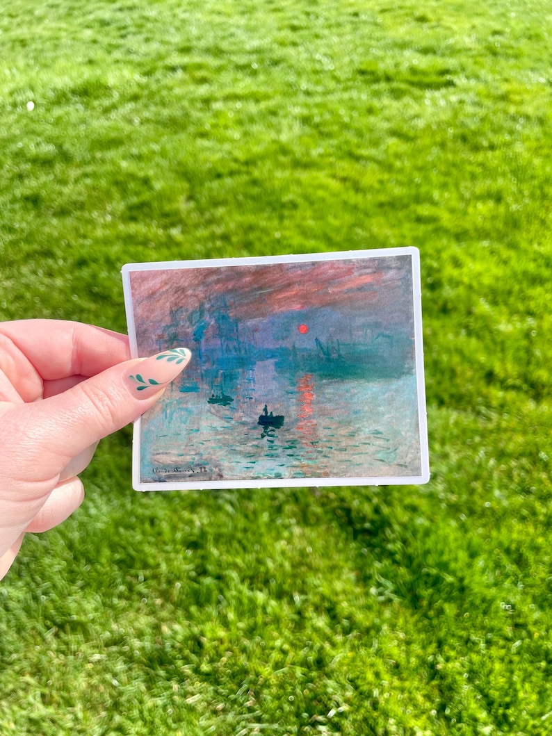 Op de afbeelding: Een sticker met een schilderij van een zonsopgang boven water met een klein bootje op de voorgrond. De lucht is een mix van roze, oranje en paars, en het water is diepblauw. Het schilderij is omlijst met een witte rand.