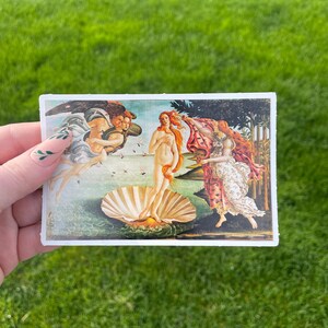 May include: A sticker featuring a reproduction of Sandro Botticelli's The Birth of Venus painting. The painting depicts a nude woman standing on a giant scallop shell, with two figures flanking her. The woman is holding her hair and looking to the left. The figure on the left is a winged figure blowing wind towards the woman. The figure on the right is a woman holding a red robe.