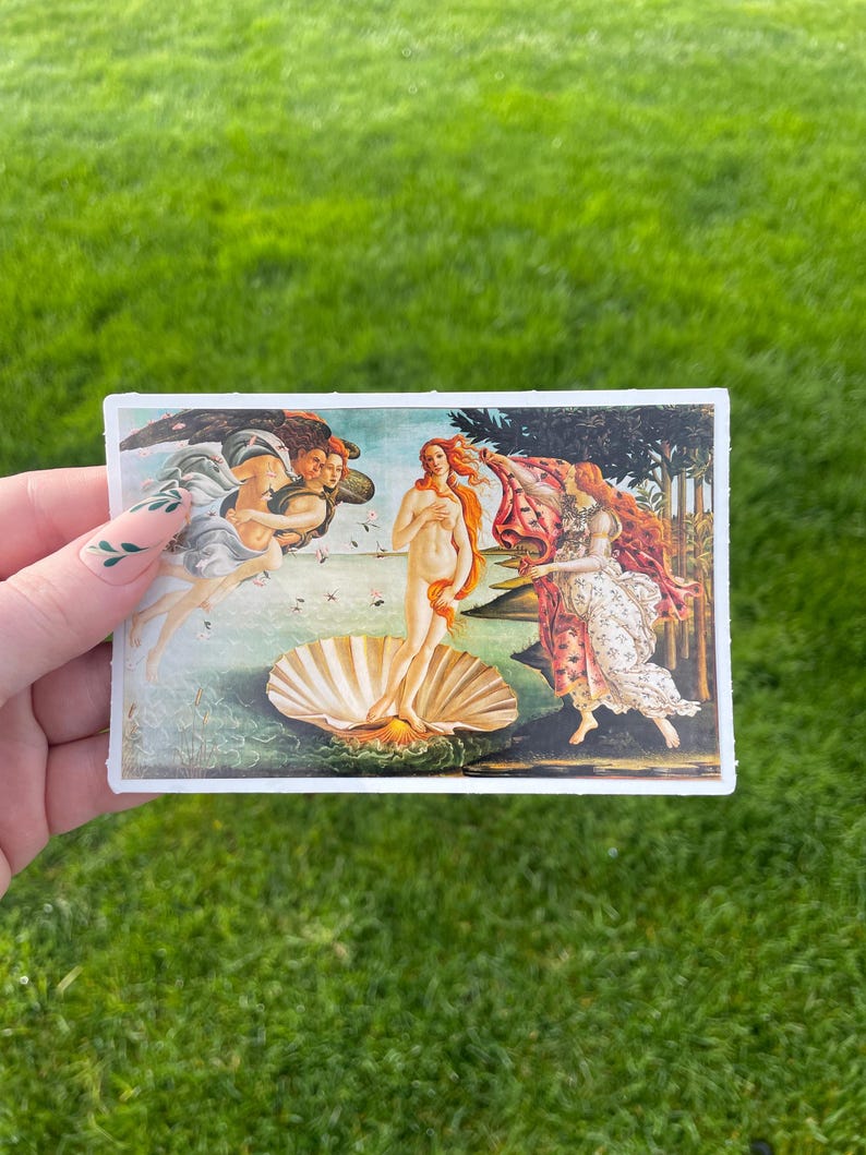 May include: A sticker featuring a reproduction of Sandro Botticelli's The Birth of Venus painting. The painting depicts a nude woman standing on a giant scallop shell, with two figures flanking her. The woman is holding her hair and looking to the left. The figure on the left is a winged figure blowing wind towards the woman. The figure on the right is a woman holding a red robe.