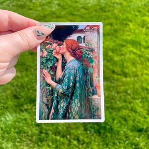 May include: A woman in a blue and gold patterned robe is smelling a pink rose. The rose is part of a bush growing against a stone wall. The woman has red hair and is wearing a gold necklace.
