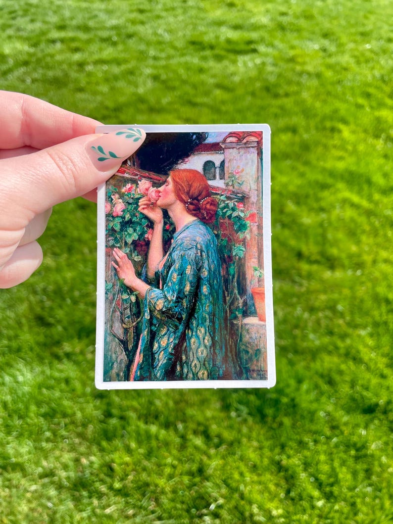 May include: A woman in a blue and gold patterned robe is smelling a pink rose. The rose is part of a bush growing against a stone wall. The woman has red hair and is wearing a gold necklace.