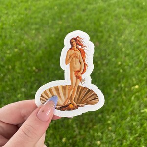 May include: A white sticker with a depiction of the Birth of Venus, a famous painting by Sandro Botticelli. The sticker features a woman standing on a seashell, with her hair flowing behind her.