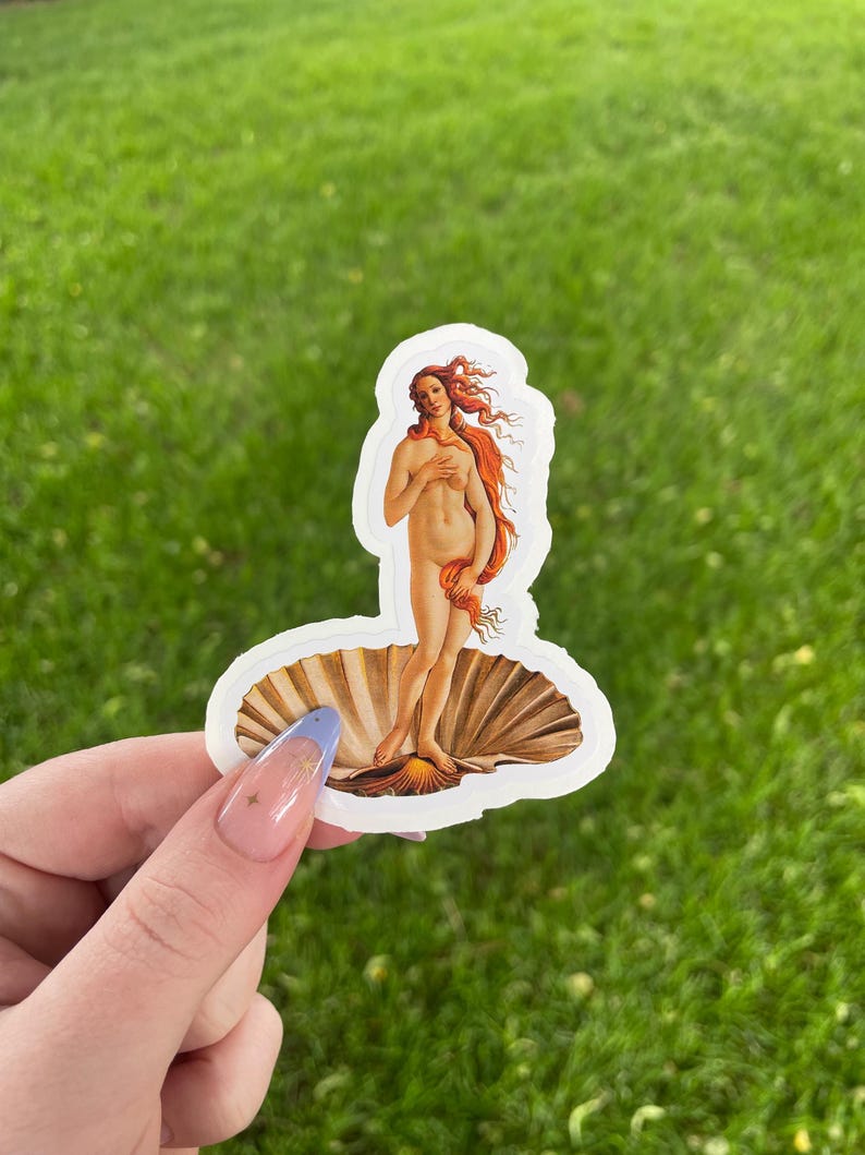 May include: A white sticker with a depiction of the Birth of Venus, a famous painting by Sandro Botticelli. The sticker features a woman standing on a seashell, with her hair flowing behind her.