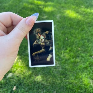 May include: A sticker featuring a painting of a grotesque figure with a large mouth and a small figure in its hand.