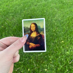 May include: A sticker featuring a reproduction of the Mona Lisa painting. The sticker is held in a hand against a green grassy background.