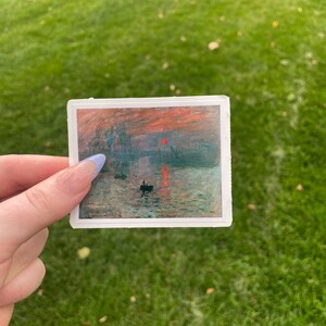 Op de afbeelding: Een sticker met een schilderij van een zeegezicht met een rode zon die ondergaat boven het water. Er zijn verschillende boten in het water en een paar gebouwen in de verte.