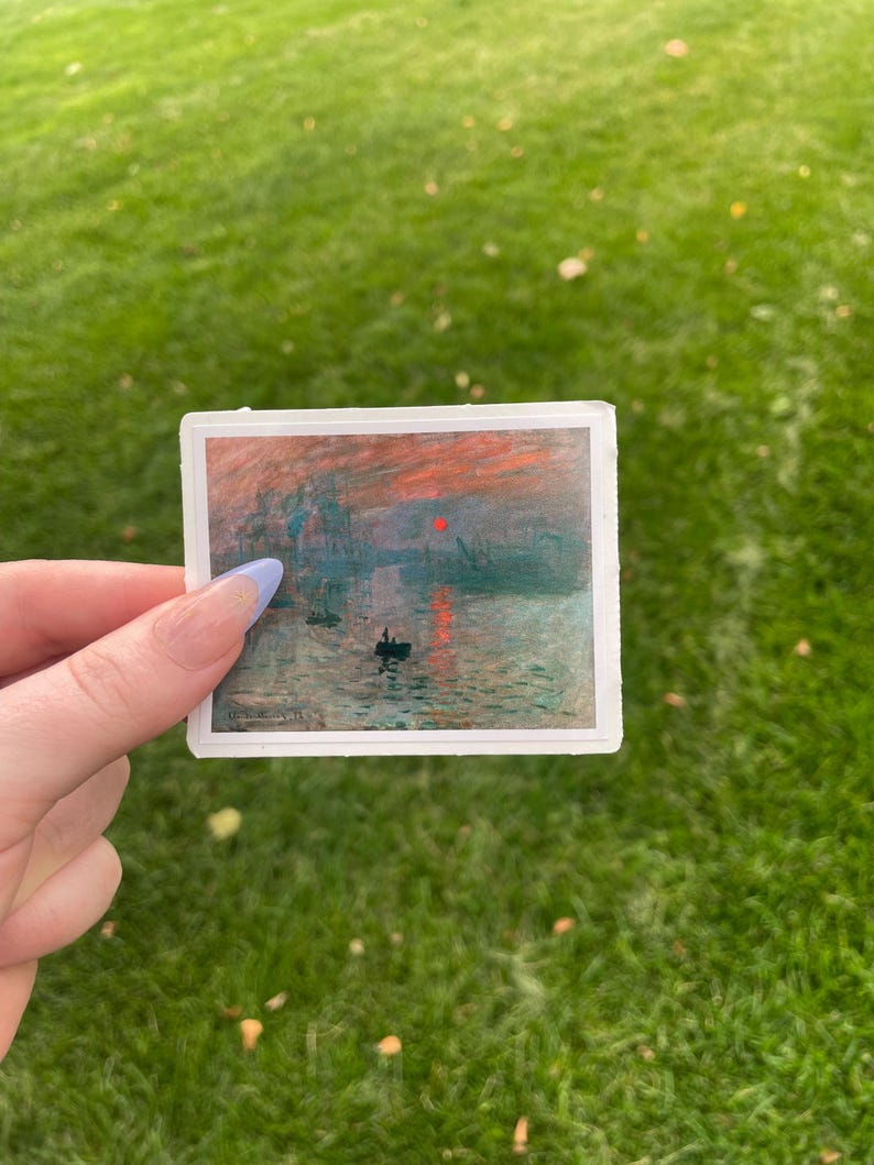 Op de afbeelding: Een sticker met een schilderij van een zeegezicht met een rode zon die ondergaat boven het water. Er zijn verschillende boten in het water en een paar gebouwen in de verte.