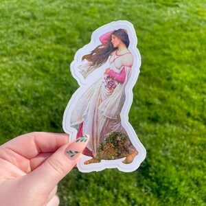 May include: A sticker featuring a vintage illustration of a woman in a white dress with pink accents, holding a bouquet of flowers. Her hair is flowing in the wind. The sticker is on a green grassy background.