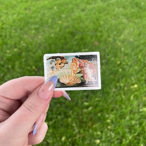 May include: A sticker featuring a reproduction of Sandro Botticelli's The Birth of Venus painting. The painting depicts a nude woman standing on a giant scallop shell, with the god of the wind, Zephyrus, and the nymph, Chloris, blowing her towards the shore.