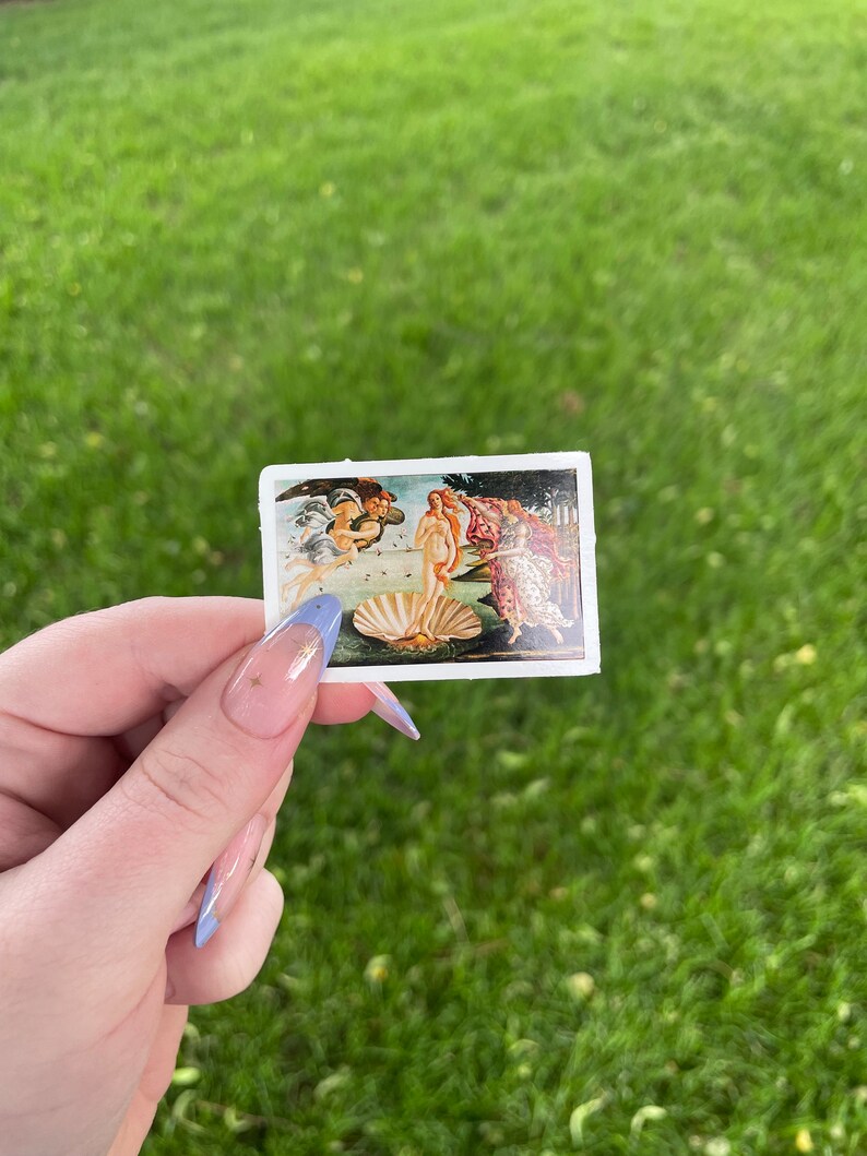 May include: A sticker featuring a reproduction of Sandro Botticelli's The Birth of Venus painting. The painting depicts a nude woman standing on a giant scallop shell, with the god of the wind, Zephyrus, and the nymph, Chloris, blowing her towards the shore.