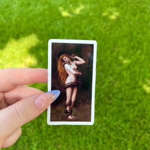 May include: A sticker featuring a painting of a woman with long red hair, holding a snake around her waist. The woman is nude and standing in a forest setting.