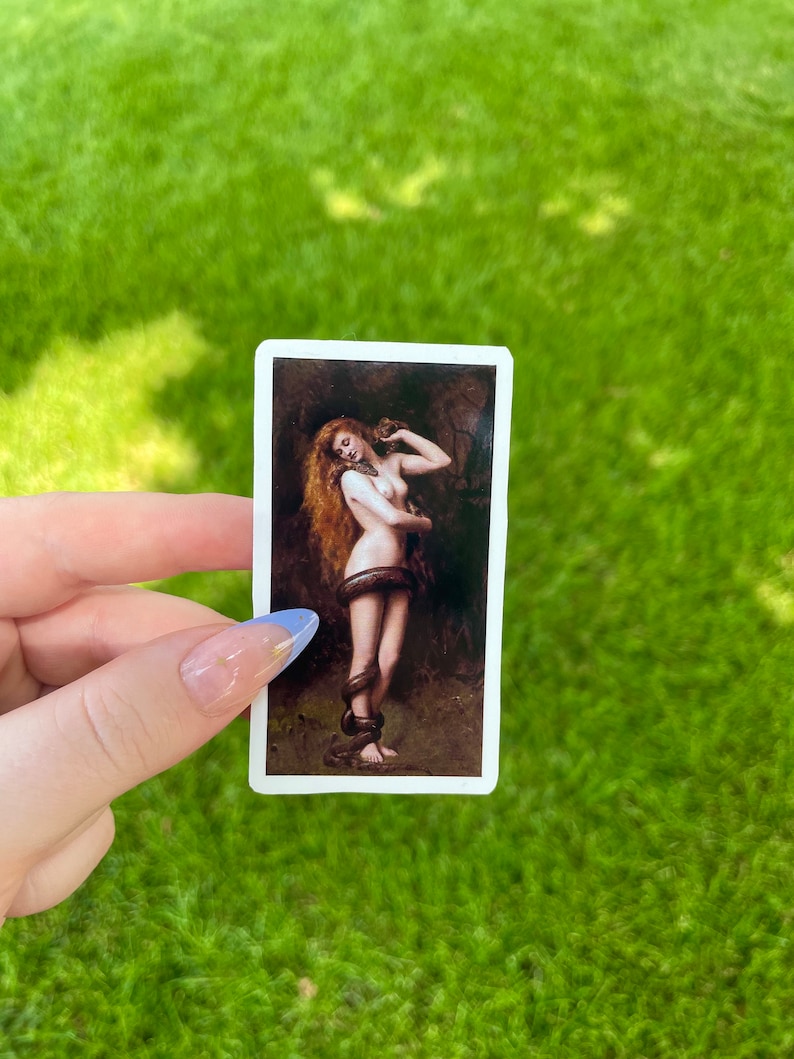 May include: A sticker featuring a painting of a woman with long red hair, holding a snake around her waist. The woman is nude and standing in a forest setting.