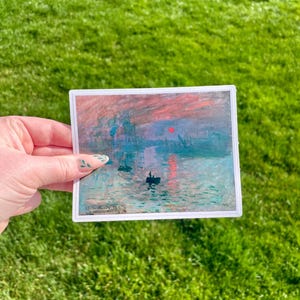 Op de afbeelding: Een sticker met een schilderij van een zonsopgang boven een wateroppervlak met verschillende schepen in de verte. De lucht is een mix van roze, oranje en blauw. Het water is lichtblauw met een reflectie van de zon.