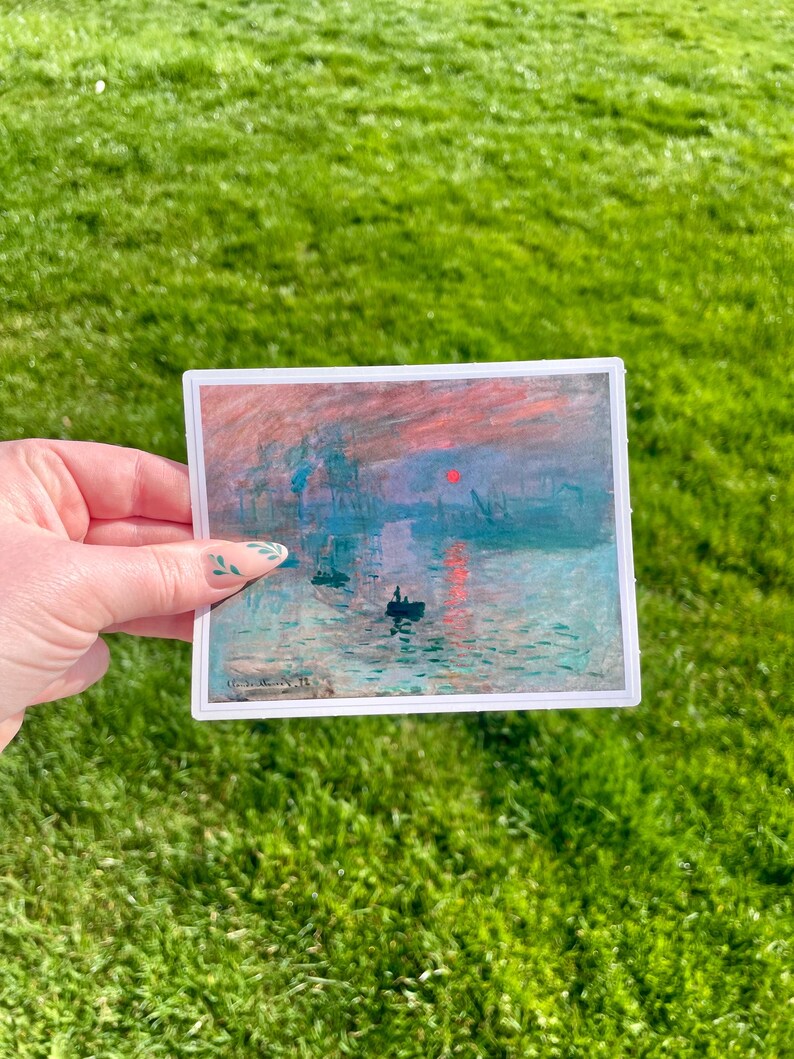Op de afbeelding: Een sticker met een schilderij van een zonsopgang boven een wateroppervlak met verschillende schepen in de verte. De lucht is een mix van roze, oranje en blauw. Het water is lichtblauw met een reflectie van de zon.