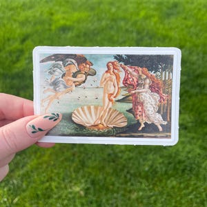 May include: A sticker featuring a reproduction of Sandro Botticelli's The Birth of Venus painting. The painting depicts a nude woman standing on a giant scallop shell, with the wind blowing her hair and a figure holding a cloak behind her.