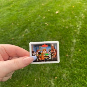 May include: A sticker featuring a painting of dogs playing cards around a green table. The dogs are wearing clothes and have human-like expressions. The painting is set in a room with a red lampshade and a clock on the wall.