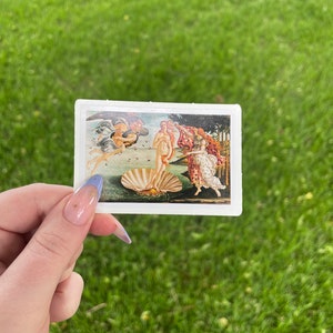 May include: A sticker featuring a reproduction of Sandro Botticelli's The Birth of Venus painting. The painting depicts a nude woman standing on a giant scallop shell, with the wind blowing her hair and a flowing robe. Two figures, one winged and one holding a cloak, are in the background.